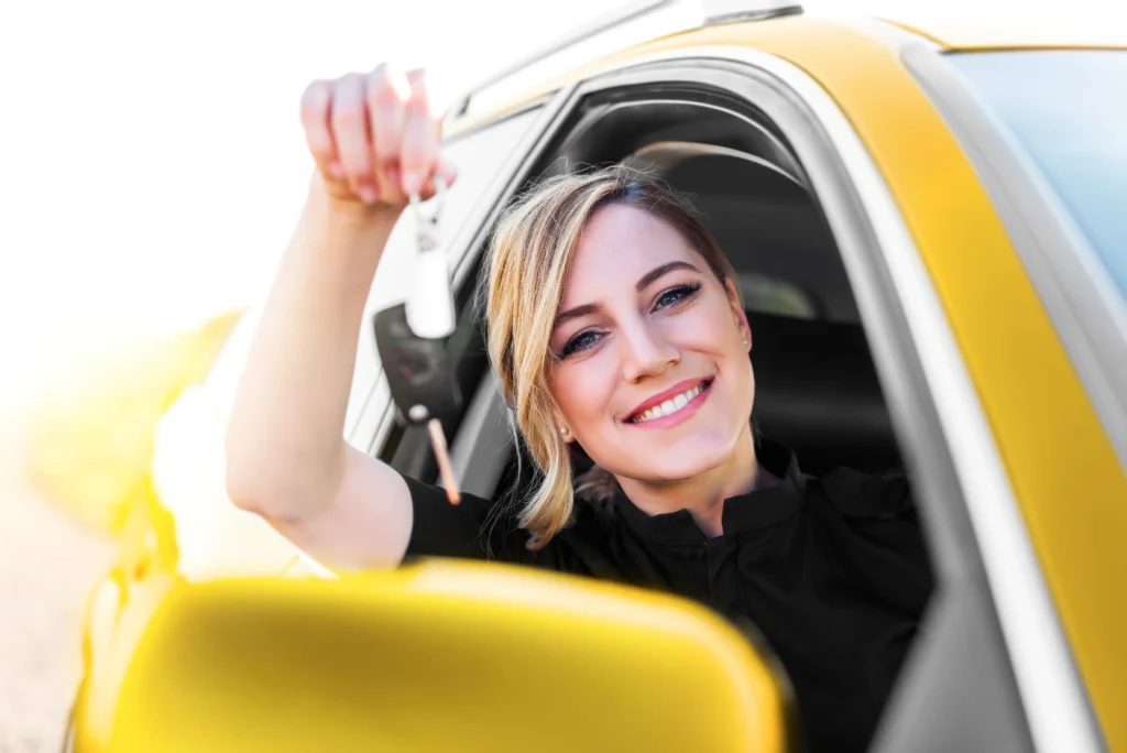 Blonde Frau lächelt aus einem gelben Auto und hält einen Autoschlüssel in der Hand